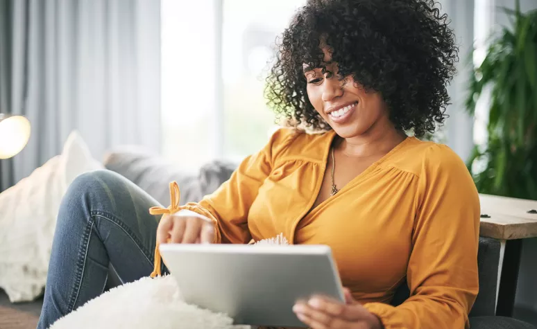 A woman smiles as she reads financial information on her tablet in her living room. A woman smiles as she reads financial information on her tablet in her living room.