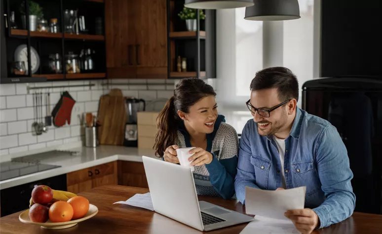 A young couple reviews retirement documents and their retirement account on their laptop in their kitchen. A young couple reviews retirement documents and their retirement account on their laptop in their kitchen.