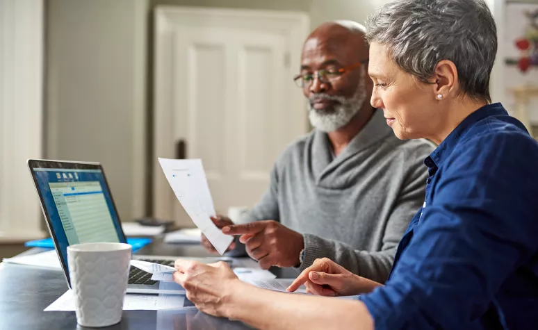 A retirement-aged couple reviews documents together in front of their open laptop. A retirement-aged couple reviews documents together in front of their open laptop.