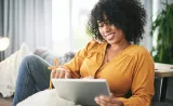 A woman smiles as she reads financial information on her tablet in her living room.