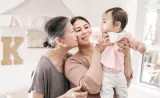 Mother smiling, holding toddler child, while grandmother looks on smiling