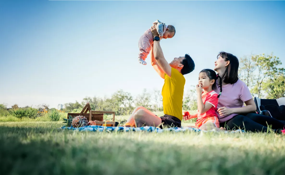 Une jeune famille pique-nique au parc. Un parent joue avec un bébé qu'il tient en l'air, tandis que l'autre est assis avec un enfant qui mange une pomme.