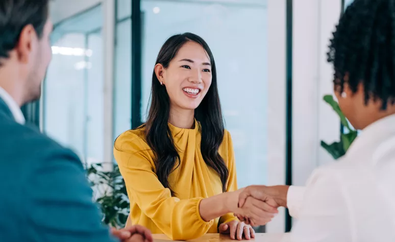 Two people shaking hands across a desk in an office, with another person seated nearby Two people shaking hands across a desk in an office, with another person seated nearby